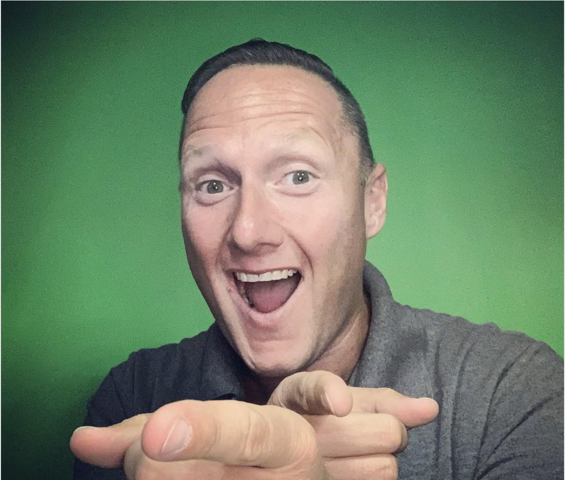 Wayne Partello smiling and pointing at the camera in a selfie against a green background.