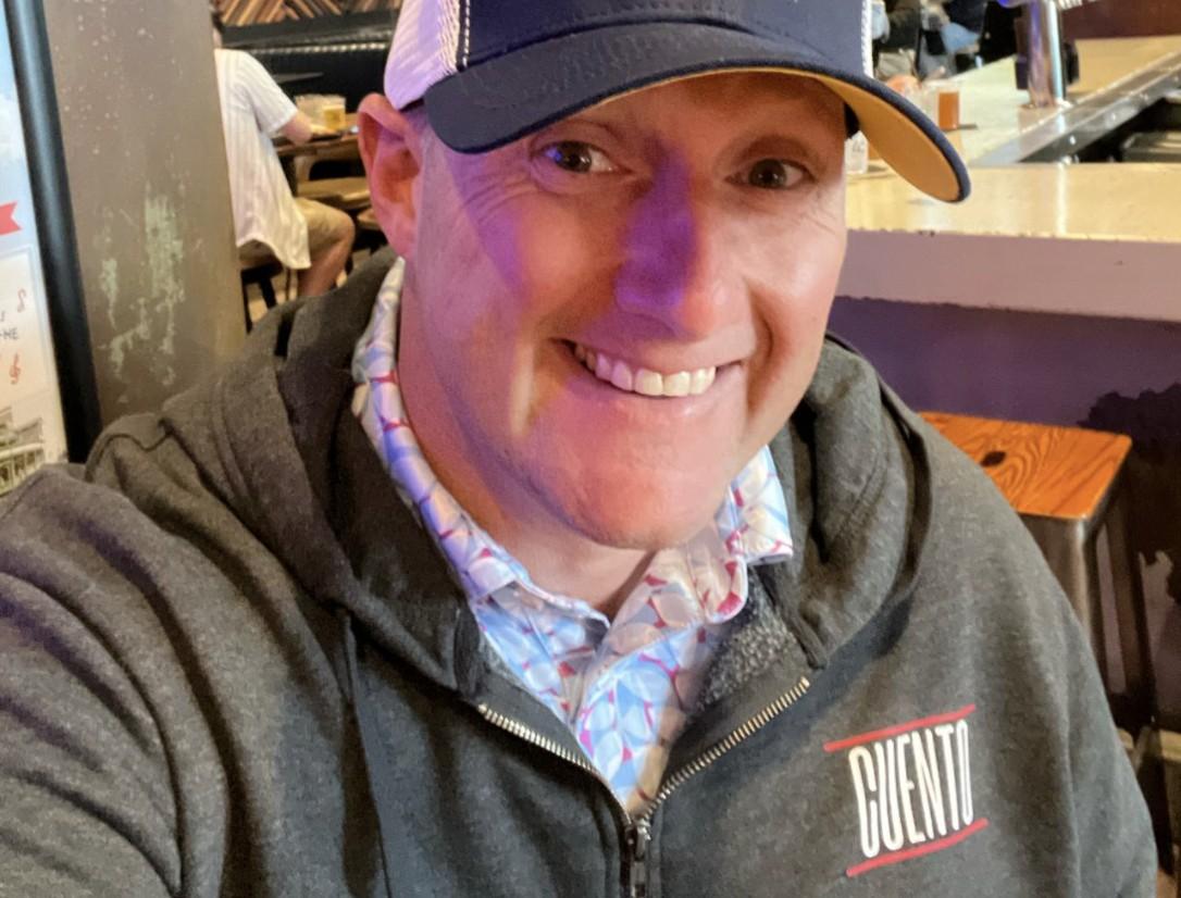 Wayne Partello smiles in a selfie wearing a Savannah Bananas cap and a CUENTO-branded hoodie.