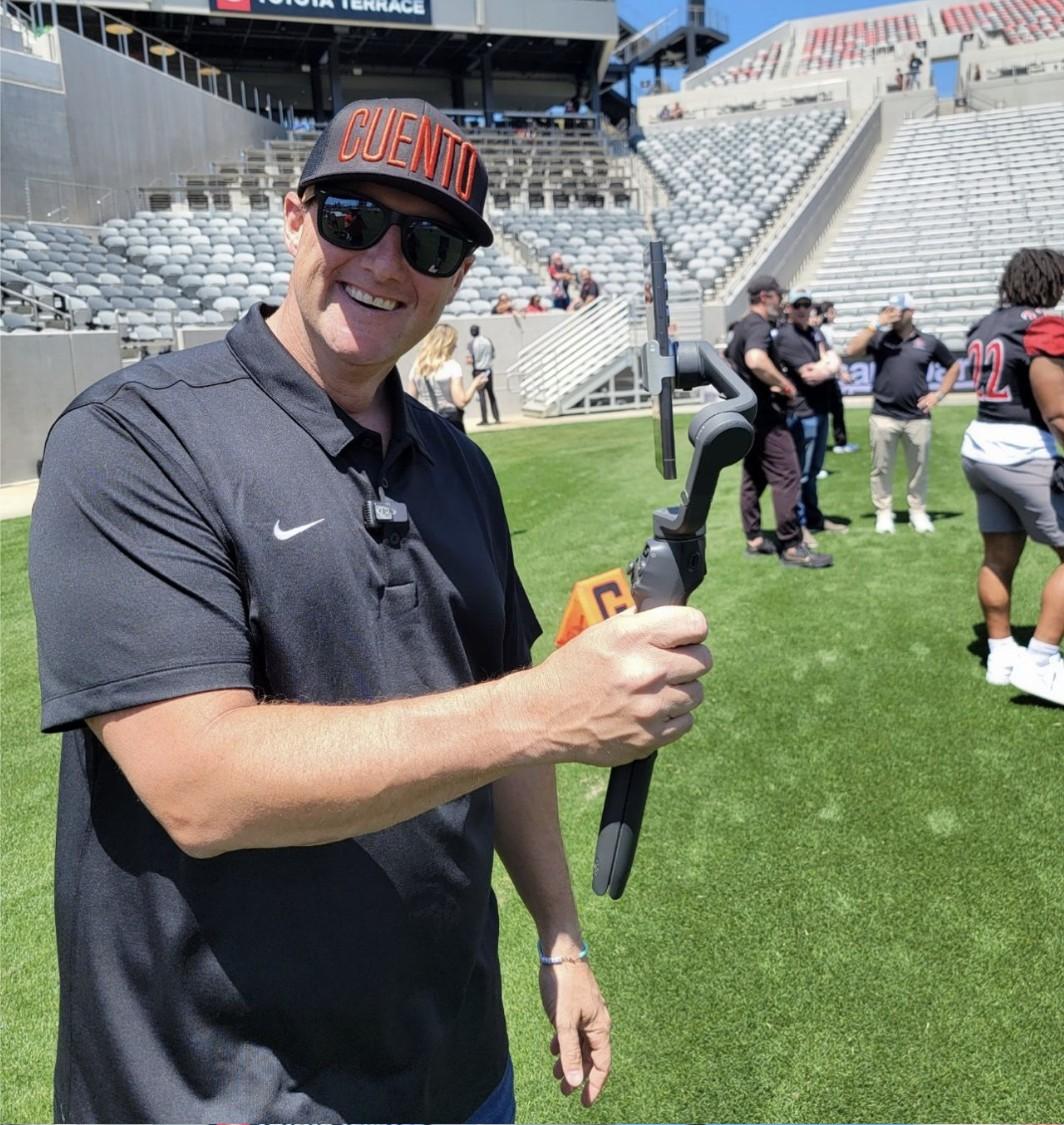 Wayne, wearing a CUENTO cap and sunglasses, holding a gimbal camera rig on the football field at a stadium during an SDSU Athletics showcase.