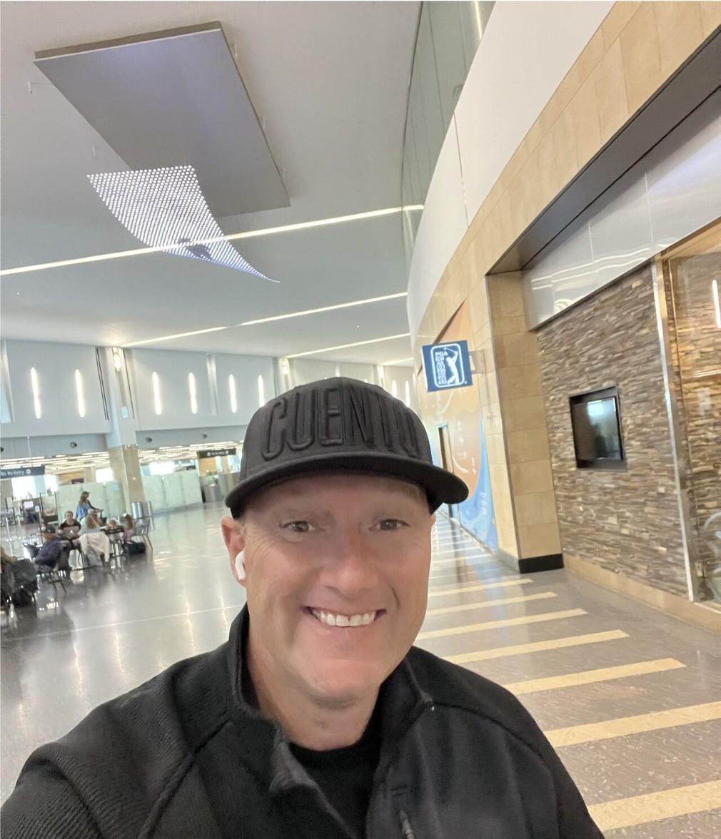 Wayne smiling in a selfie at an airport terminal, wearing a black CUENTO cap.