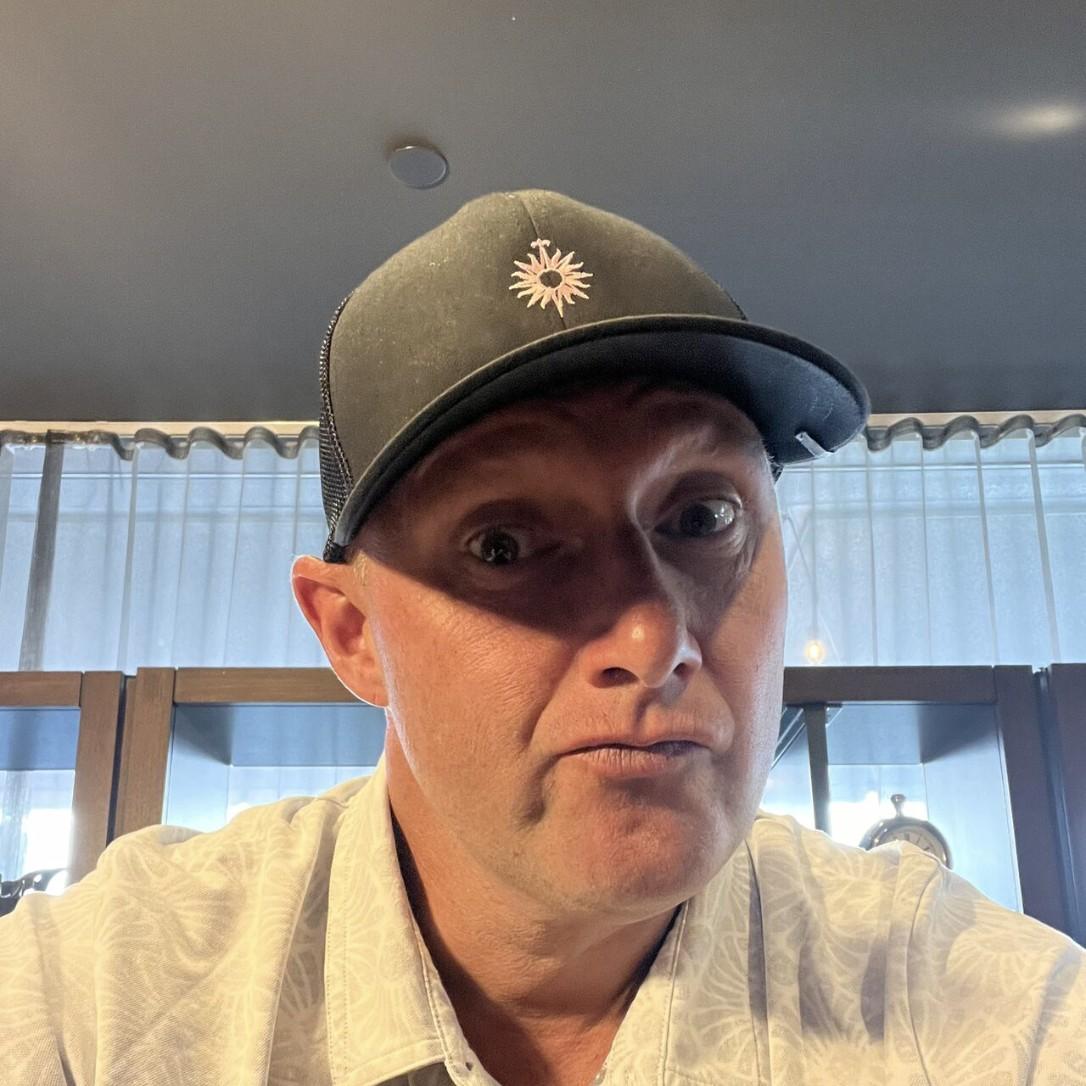 Close-up selfie of Wayne wearing a dark cap with a sunburst logo, making a skeptical expression.