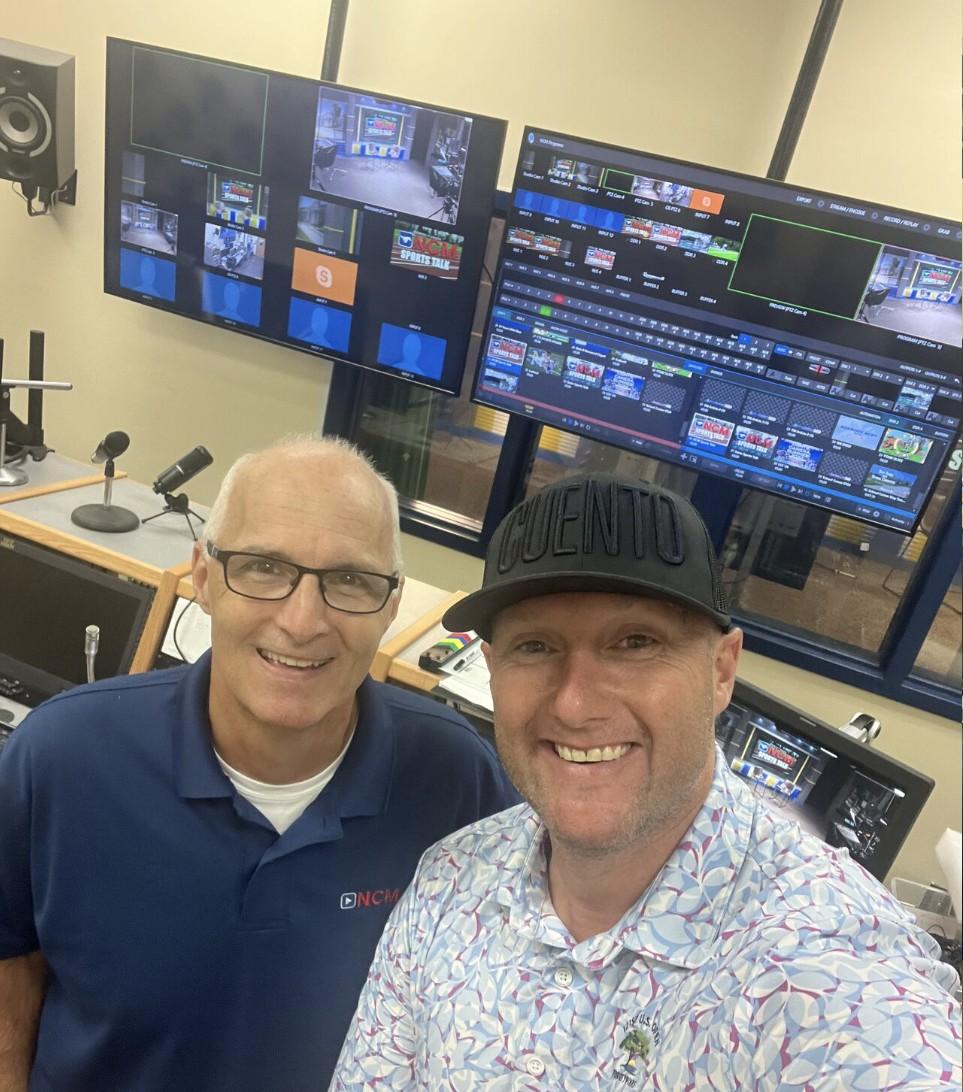 Wayne, wearing a CUENTO cap, smiling with his former teacher in a television broadcast control room full of monitors.