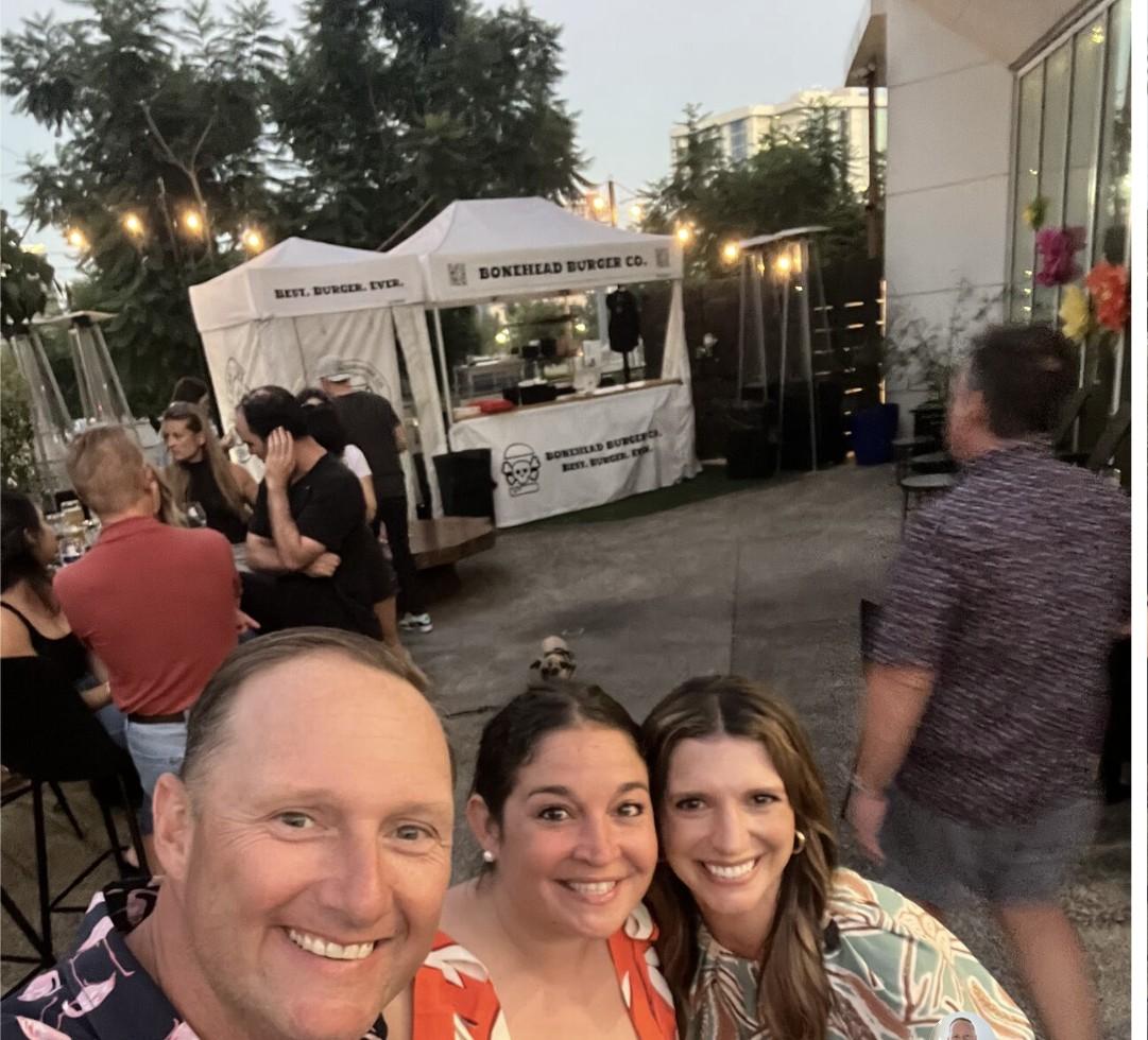 Wayne taking a selfie with two colleagues at an outdoor evening event with string lights and a food truck in the background.