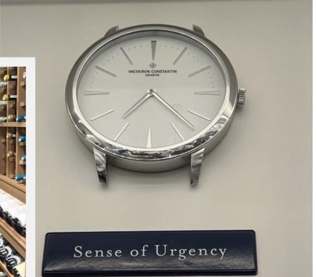 A large decorative wall clock styled like a wristwatch, labeled 'Sense of Urgency,' mounted above a wine cellar display.