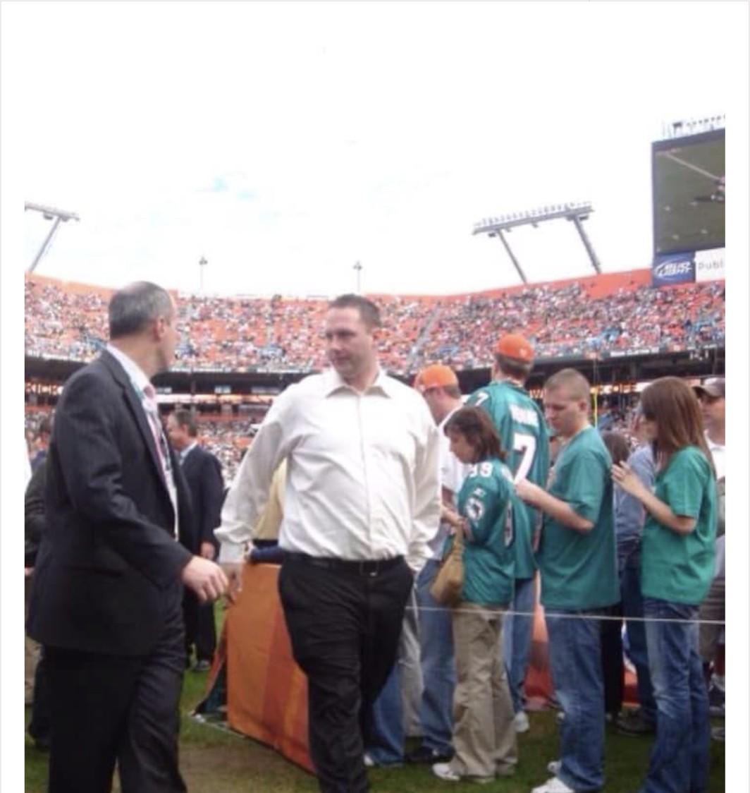 Vintage photo of a young Wayne Partello walking on the field at a packed Miami Dolphins stadium alongside a colleague in a suit, with fans in Dolphins jerseys nearby