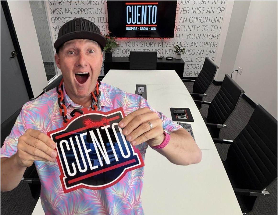 Wayne Partello smiling and holding up a CUENTO logo sign in the CUENTO office conference room, with a screen displaying 'CUENTO: Inspire, Grow, Win' in the background