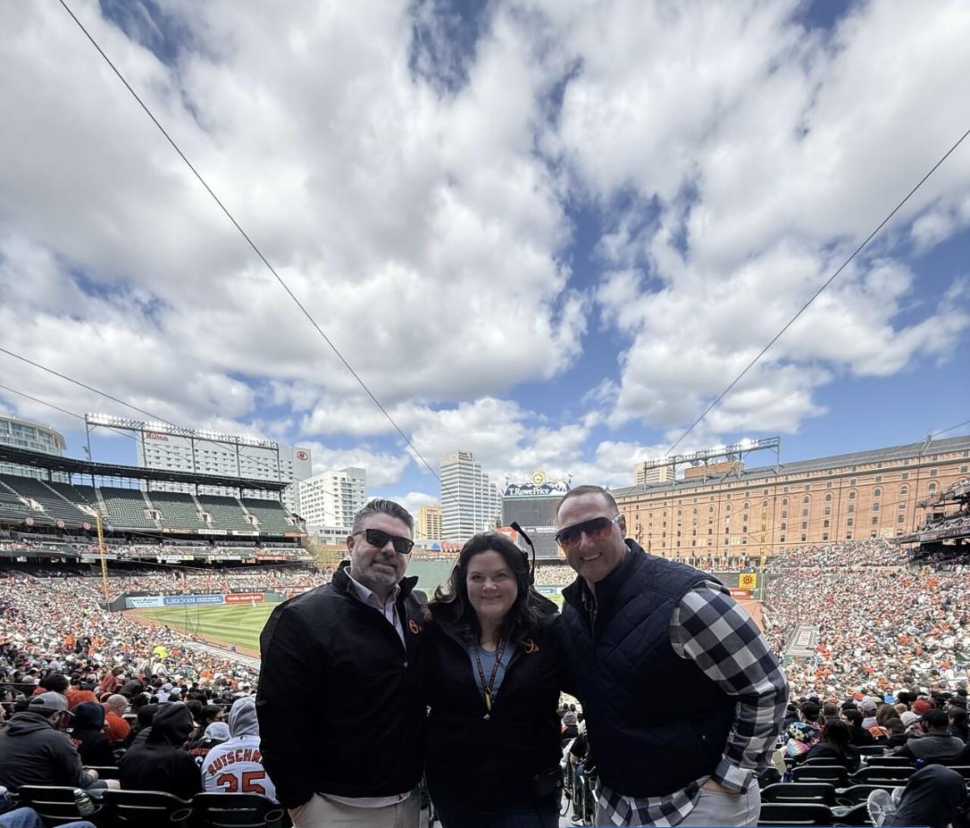 Wayne Partello and two colleagues smiling together in the stands at Camden Yards, Baltimore Orioles stadium, under a partly cloudy sky