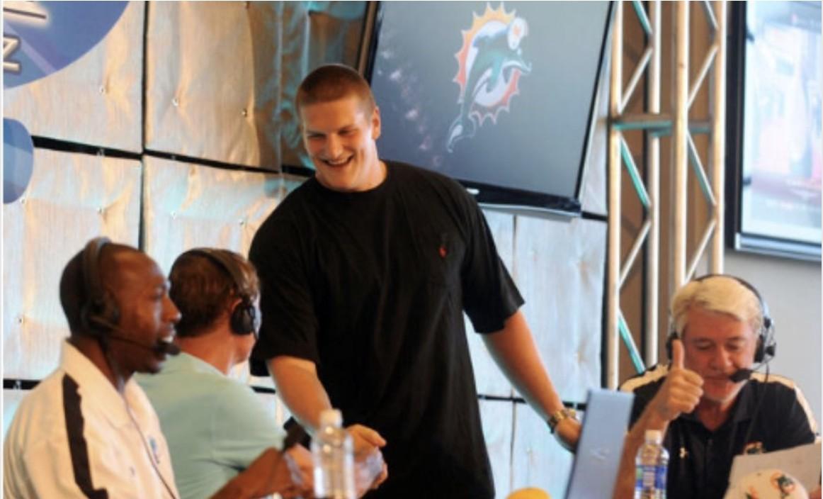 A young Miami Dolphins draft pick smiling and shaking hands on stage with broadcast hosts wearing headsets, a Dolphins logo displayed on a screen behind them
