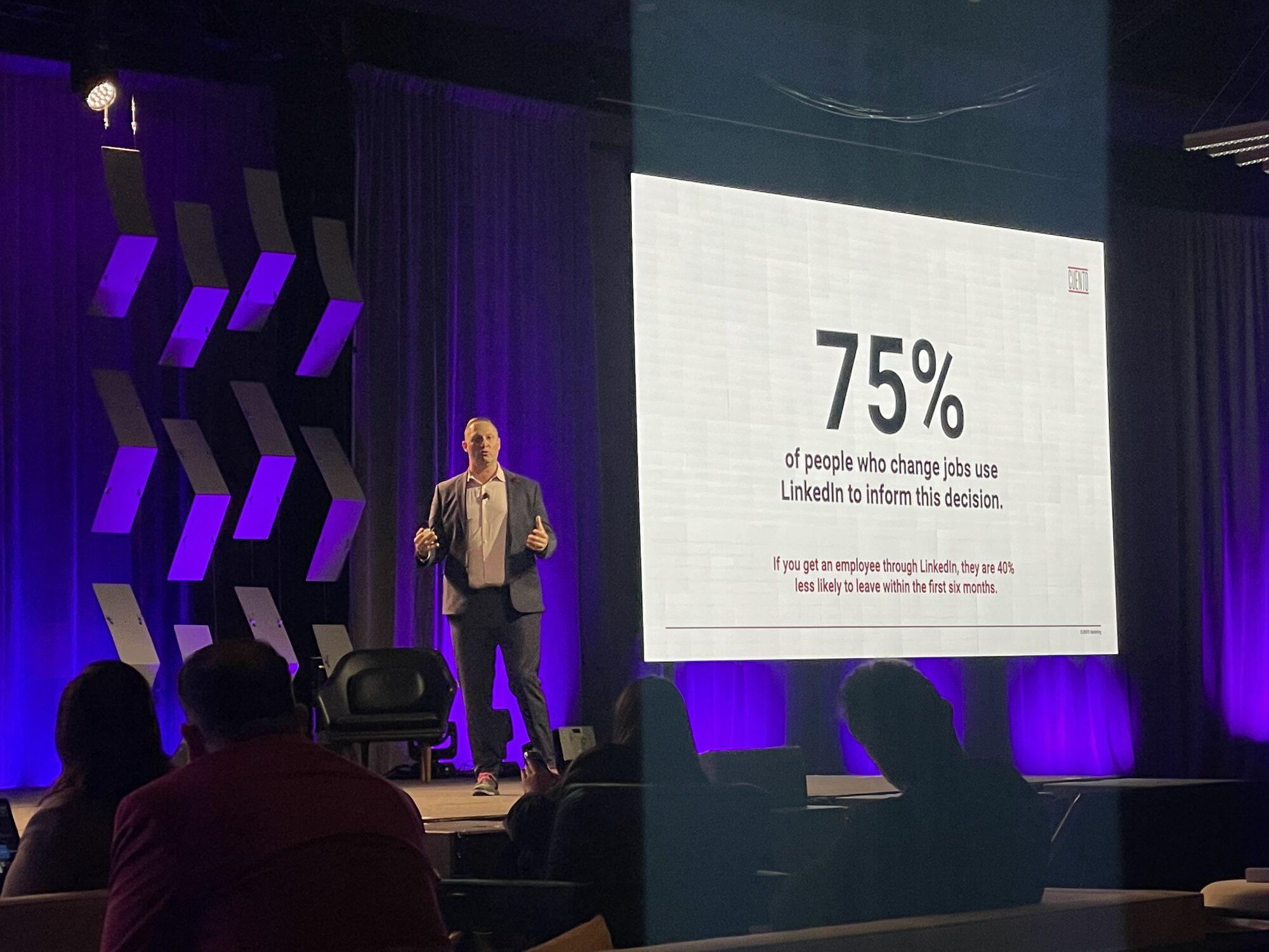 Wayne Partello speaking on stage beside a presentation slide