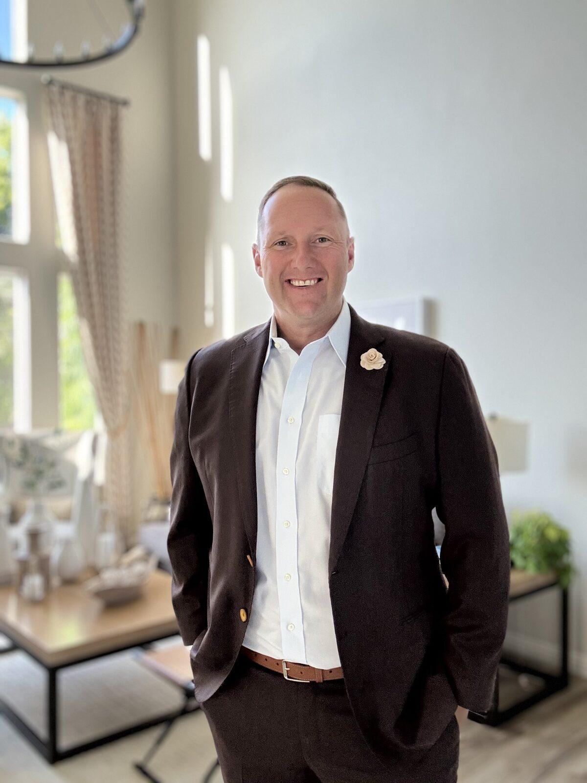 Wayne Partello in a dark blazer and open-collar white shirt, smiling, in a bright room